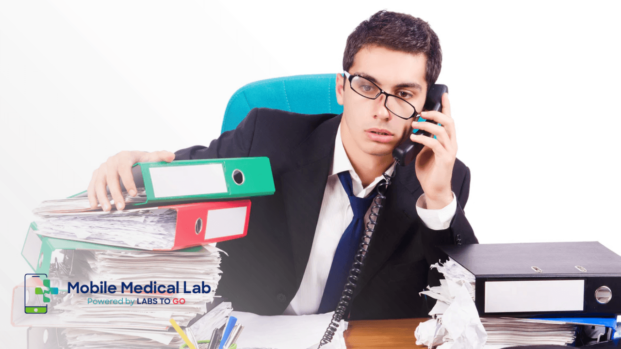 Overwhelmed professional on the phone surrounded by stacks of paperwork, representing the logistics that cause people to delay lab work